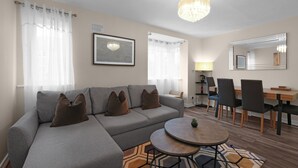 A modern living room with a grey sofa, a round coffee table, and a dining area with chairs and a table. - Modern 1 Bedroom Apt in Streatham with Parking (London)