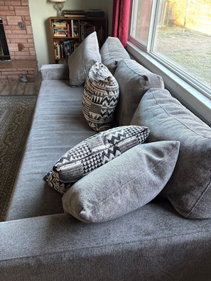A grey sofa with patterned cushions, a bookshelf, and a window with a view of a garden. - Quiet Meridian Cottage (Boise)