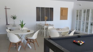 Ett modernt rum med ett biljardbord, vita stolar och ett matbord med stolar. På väggen hänger abstrakta målningar och på matbordet står en växt.