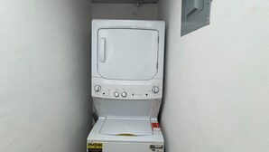 A compact, white laundry room with a front-loading washer and dryer, a metal vent, and a small electrical outlet on the wall. - Apartment 10 minutes from Las Americas International Airport and the beach. (Santo Domingo Este)