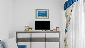 Una sala de estar con un televisor sobre un mueble blanco, un sofá azul, un cuadro enmarcado y una cortina.