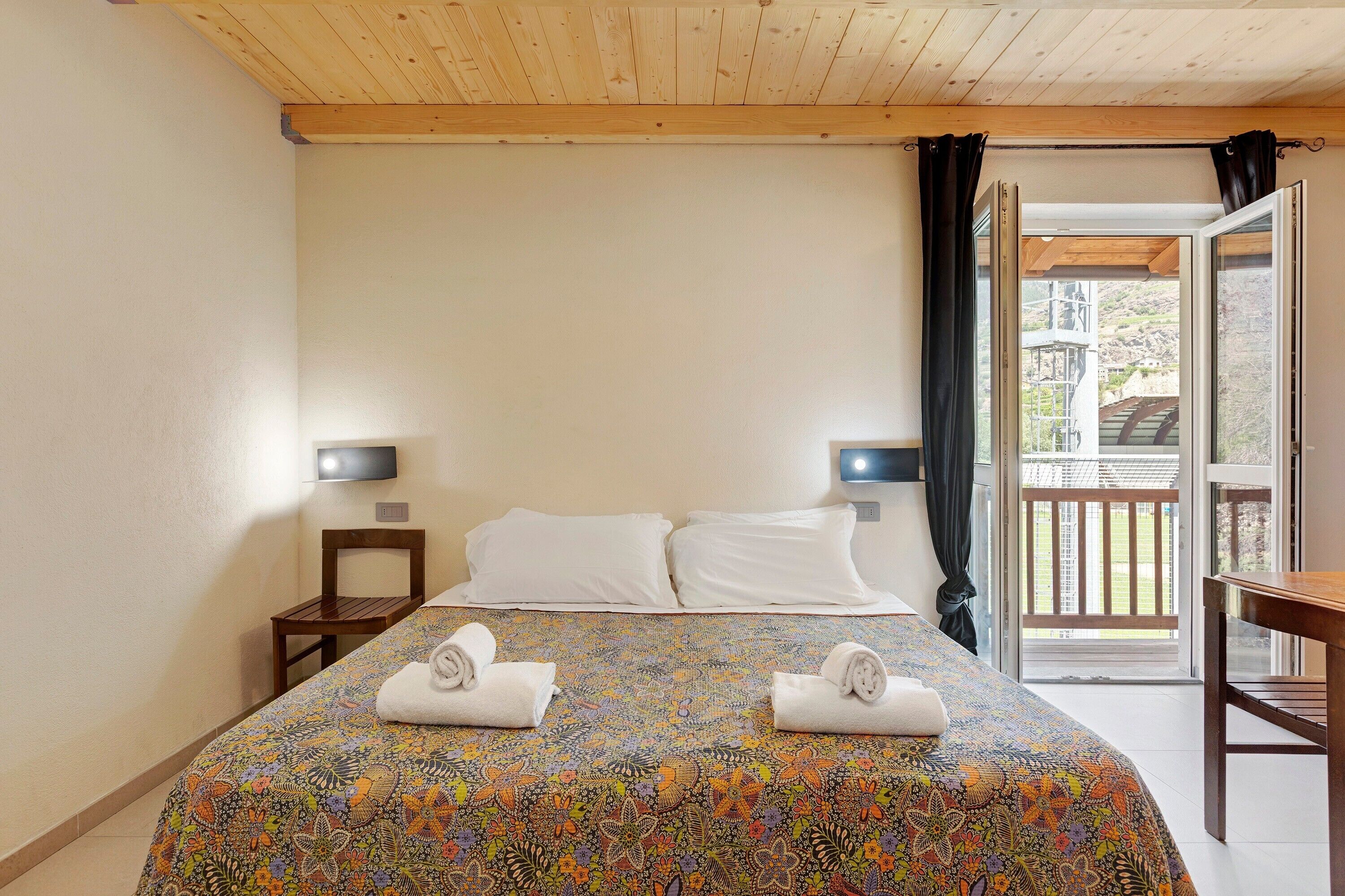 A hotel room with a bed, two bedside tables, a TV, and a balcony with a view.