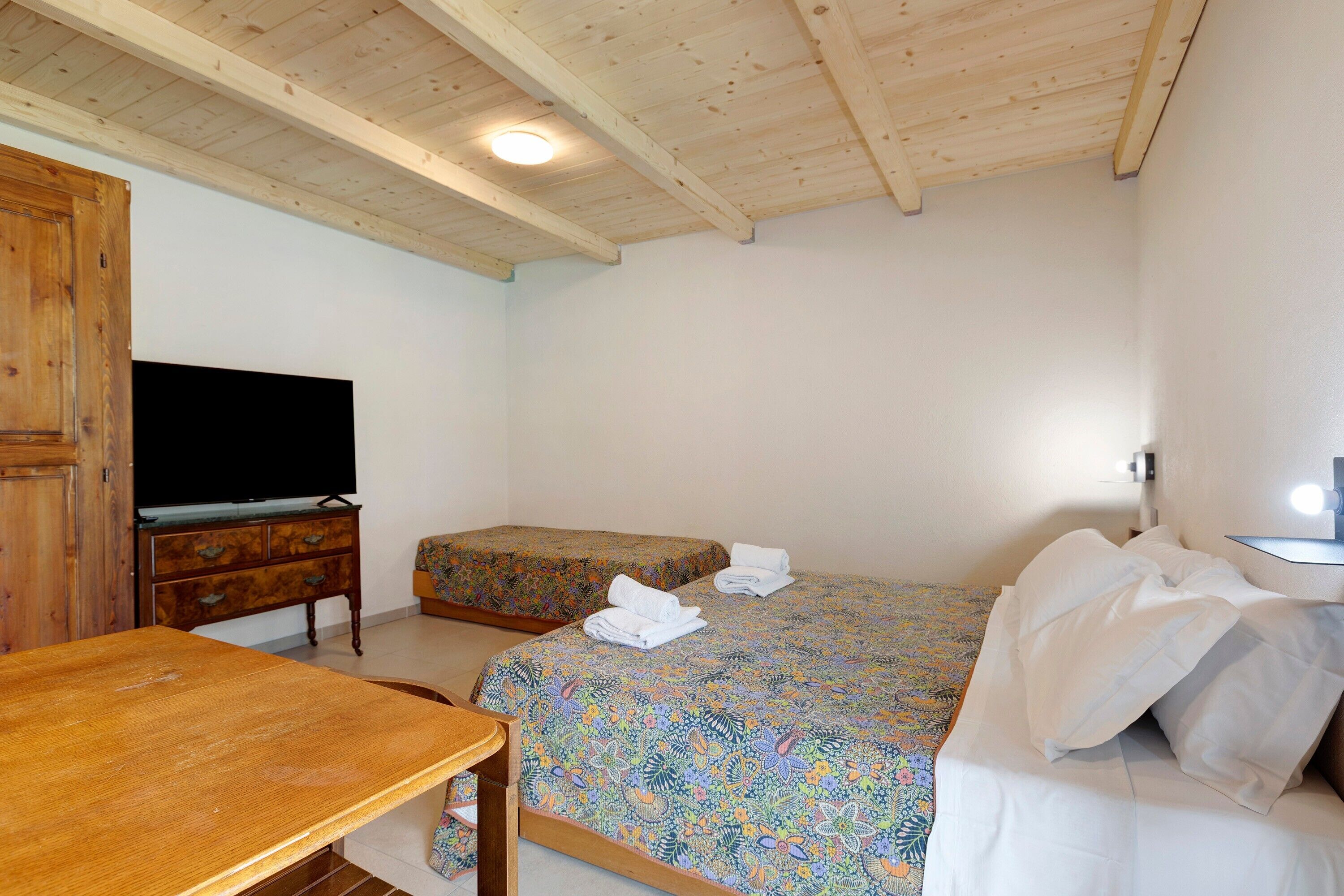 A room with a wooden ceiling, a television on a wooden cabinet, a bed with a floral bedspread, and a small table.