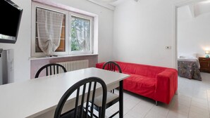 Une pièce comprenant une table à manger, des chaises, un canapé rouge, un téléviseur et une fenêtre avec des stores.