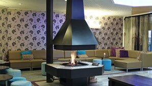 A modern lounge area with a central fire feature, sofas with patterned cushions, and a patterned wall. - A Place You'll Surely Enjoy! Indoor Pool, Near Henriette Polak Museum! (Zutphen)