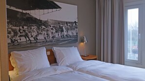 Ein Hotelzimmer mit Bett, weißen Kissen, einem Nachttisch mit Lampe, einem großen Schwarz-Weiß-Foto an der Wand und einem Fenster mit Vorhängen.