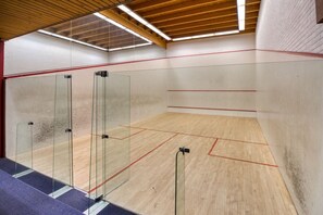 Un court de squash doté de portes en verre, d’un plancher en bois et d’un plafond incliné.