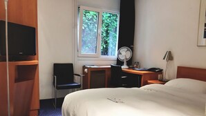 Habitación de hotel con cama, escritorio, silla, televisión, ventana con vistas a la vegetación y una lámpara de pie.