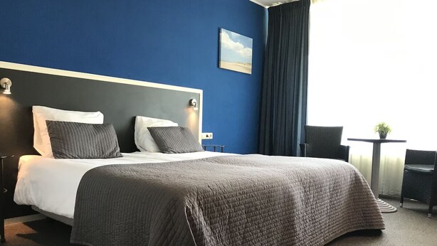 Ein Hotelzimmer mit Bett, Sessel, kleinem Tisch und einem Fenster mit Vorhängen.