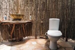 Un baño rústico con suelo de piedra, inodoro blanco, lavabo de madera y pared de bambú.