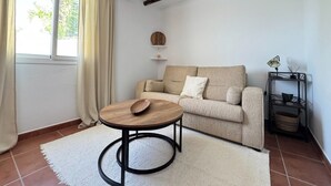 A cozy living room with a sofa, a round table, a hanging light fixture, and a window with curtains. - Cozy house in a picturesque white mountain village (Canillas de Albaida)