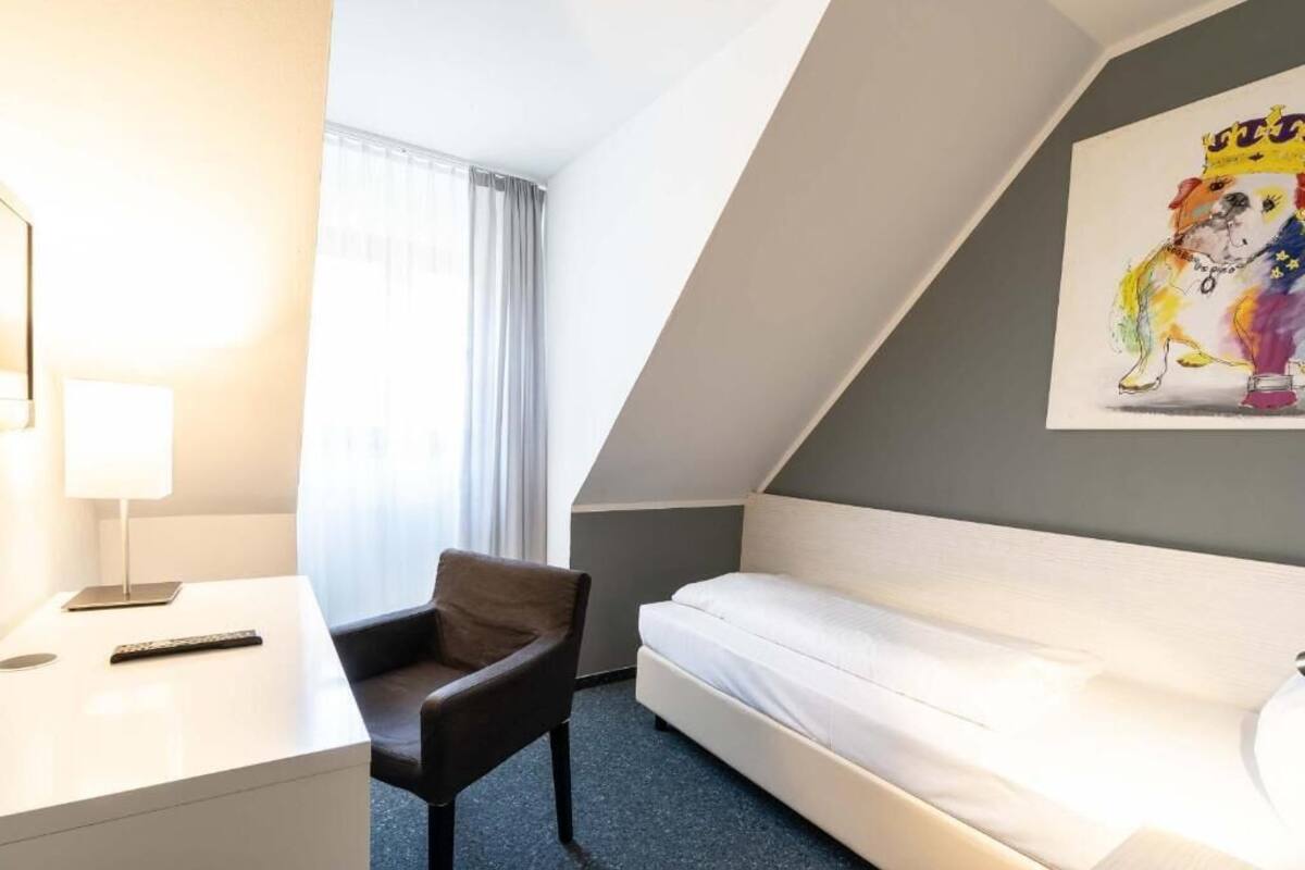 A hotel room with a bed, a desk, a chair, a window with curtains, and a painting on the wall.