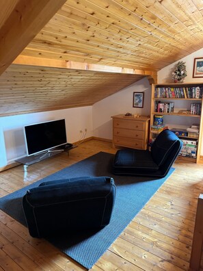 A loft bedroom with a sloped ceiling, a flat-screen TV, a bookshelf, and two black chairs on a blue mat. - Stunning Alpine chalet for seven people with breathtaking mountain views! (La Chapelle-d'Abondance)