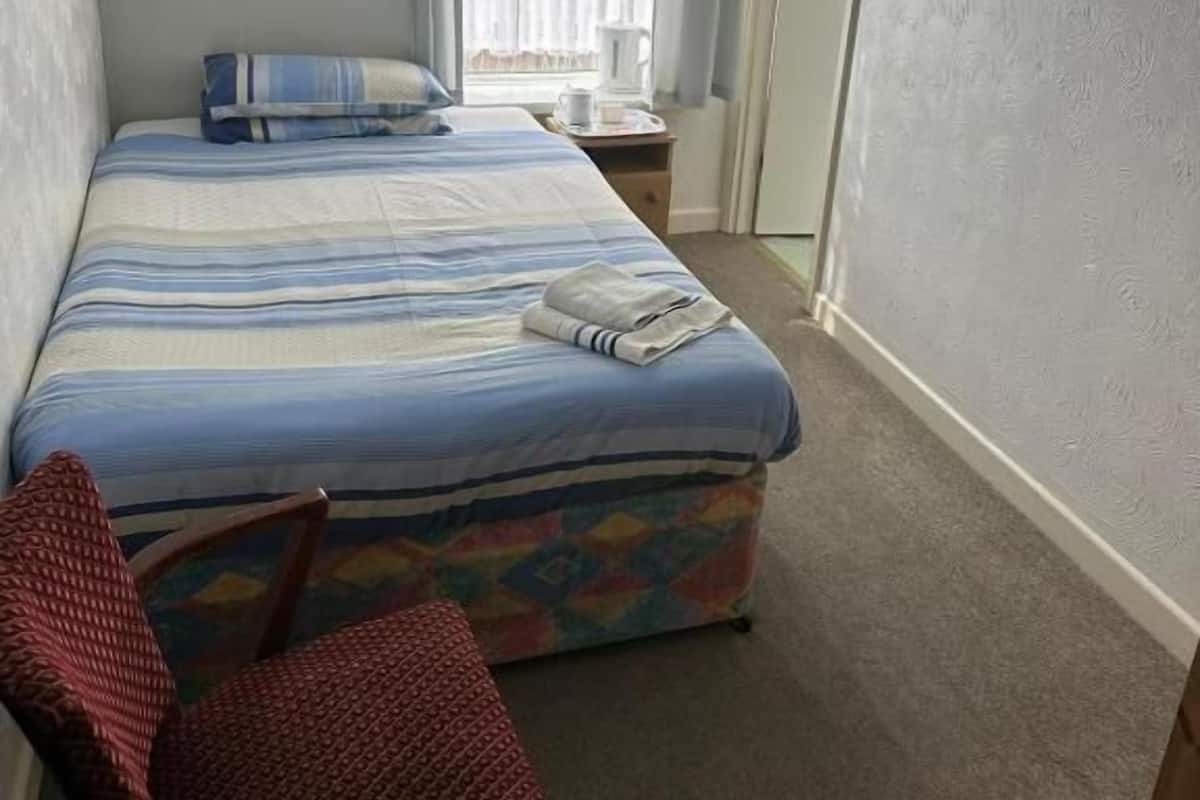 Een slaapkamer met een bed, een stoel, een raam met gordijnen, een schilderij aan de muur en een deur.