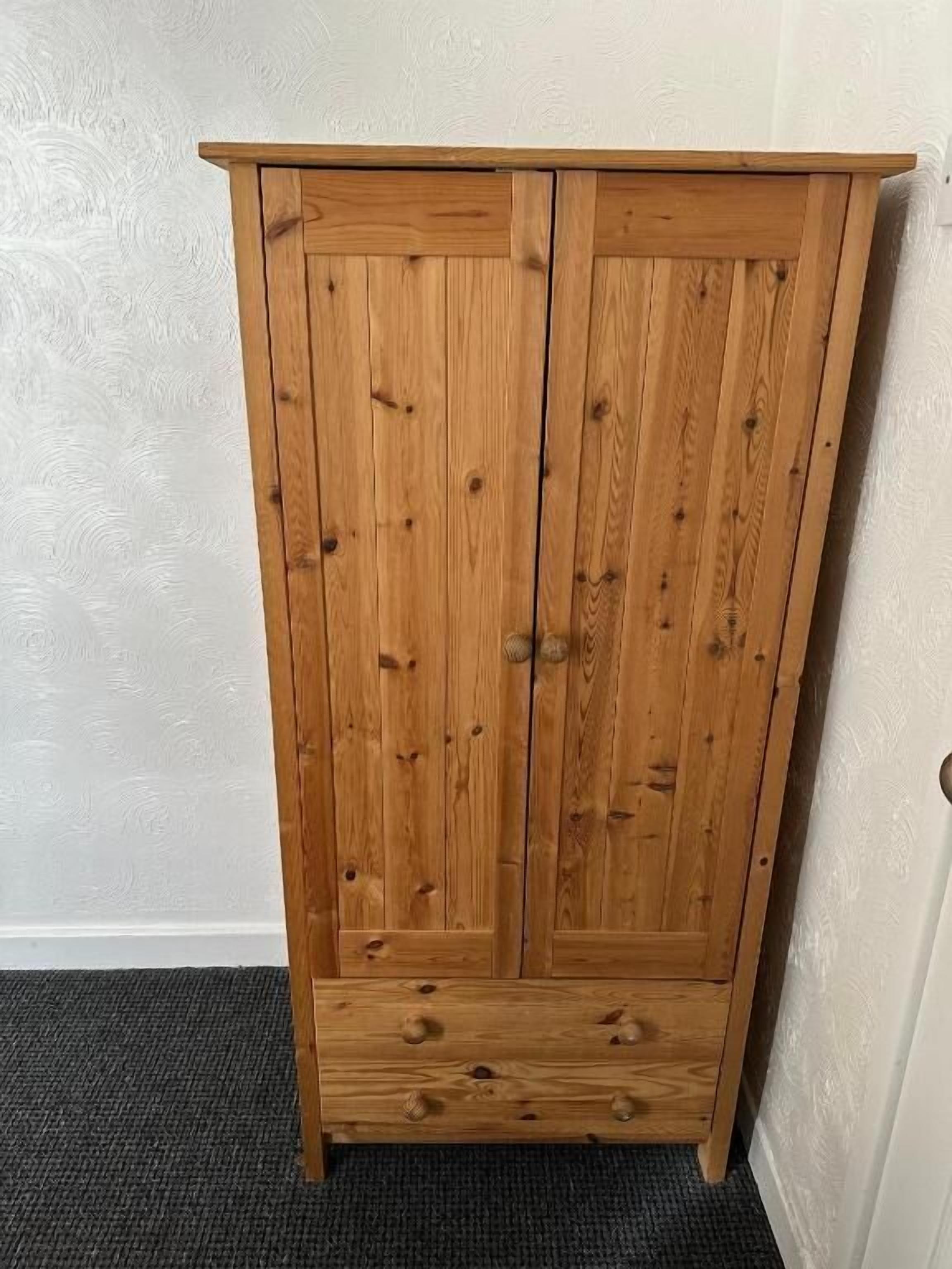 Une armoire en bois à deux portes, avec une étagère inférieure, placée contre un mur à texture.