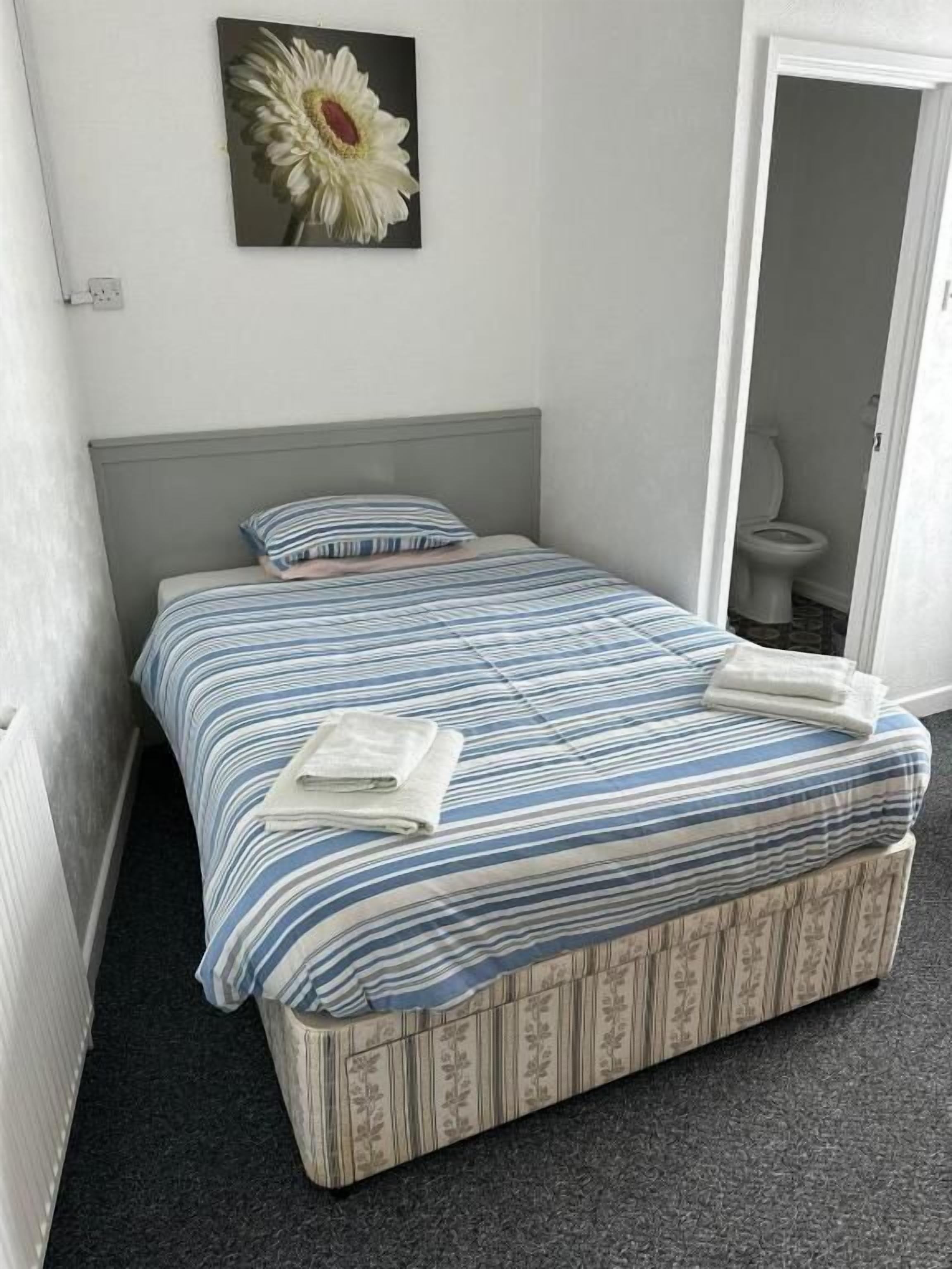 A single bed with a striped bedspread, a pillow, and a folded towel. There is a painting of a flower on the wall.