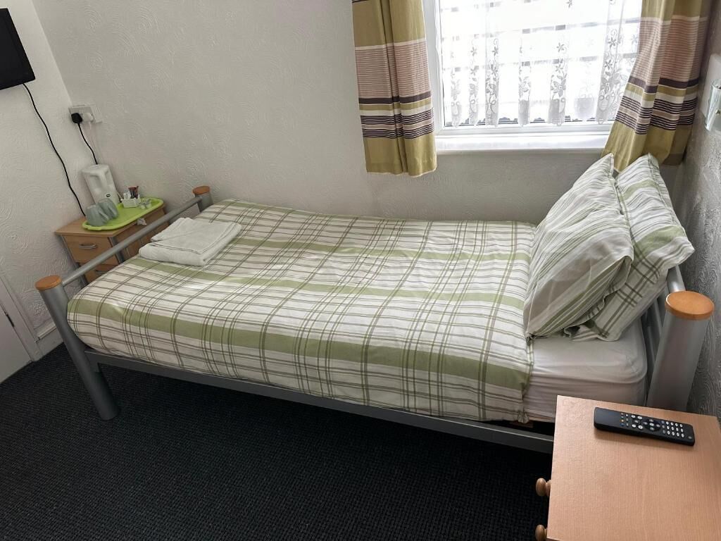 A single bed with a plaid bedspread, a bedside table with a lamp, a remote control on a small table, and a window with curtains.