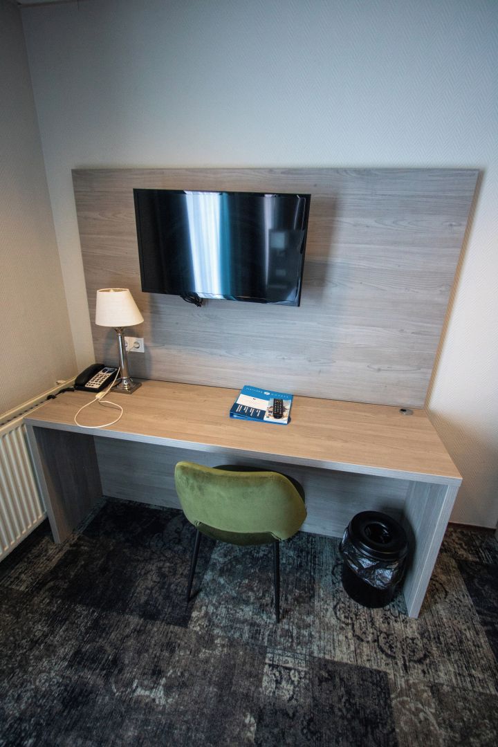 A hotel room with a desk, a television mounted on the wall, a lamp, a telephone, a chair, and a trash bin.