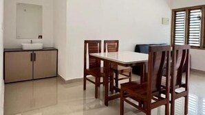A spacious room with a dining table, chairs, a ceiling fan, a window with blinds, and a kitchenette area. - Easy Nest Residency (Kannur)