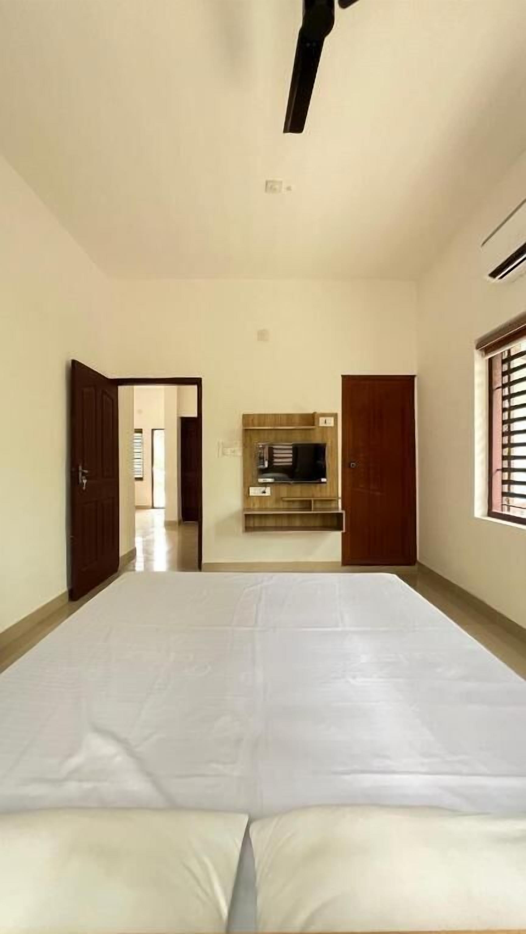 A room with a bed, a ceiling fan, a TV mounted on the wall, and a window with blinds.
