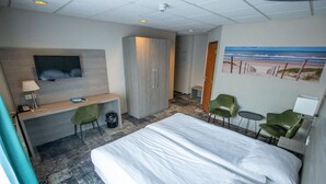 Habitación de hotel con una cama, un escritorio, dos sillas, un televisor y un cuadro de una playa.