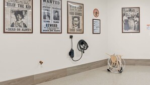 Une pièce avec un mur blanc orné de divers affiches encadrées, une prise électrique avec un câble, et une chaise en bois avec une corde.