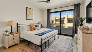 Une chambre à coucher comprenant un lit, des tables de chevet, une commode, un ventilateur de plafond et une grande fenêtre avec des rideaux.