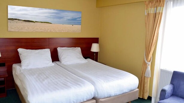 A hotel room with two beds, a wooden headboard, a painting of a beach, a window with curtains, and a blue armchair. - Family-Friendly Getaway | Near Scenic Dunes, Nature Sites, and Outdoor Trails (Bergen)