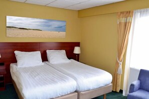Habitación de hotel con dos camas, cabecero de madera, un cuadro de playa, una ventana con cortinas y un sillón azul.