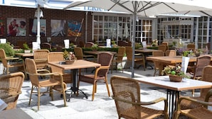 Een terras met rieten meubels, witte parasols en een bakstenen muur met kunstwerken.