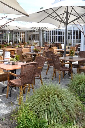 Un café en plein air avec des tables et des chaises en osier, abrité par de grands parasols et entouré de plantes en pot.