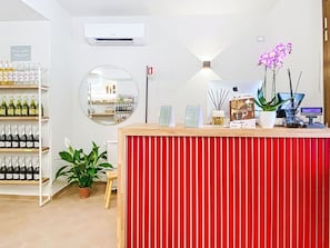 A reception desk with a red panel, a mirror, a wall-mounted air conditioner, and shelves with bottles.