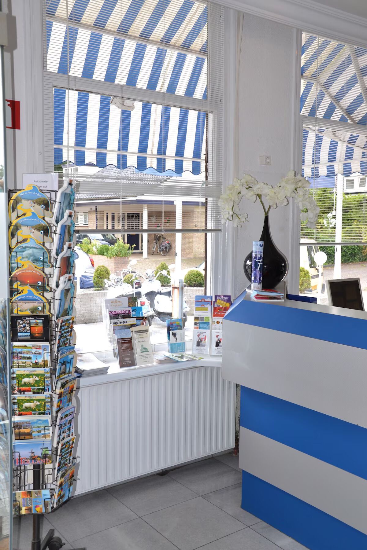 Un espace d’accueil avec un comptoir rayé de bleu et de blanc, un vase blanc contenant des fleurs blanches et un présentoir rempli de brochures de voyage.