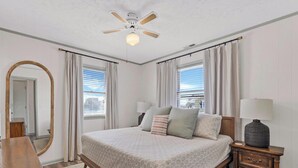 A bedroom with a bed, a nightstand, a lamp, a ceiling fan, and a window with curtains. - Spread'N Kindness | Pet-Friendly + One Block from the Beach (Surf City)