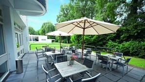 Eine Terrasse im Freien mit Tischen, Stühlen und Sonnenschirmen, mit Blick auf eine grüne Rasenfläche und Bäume.