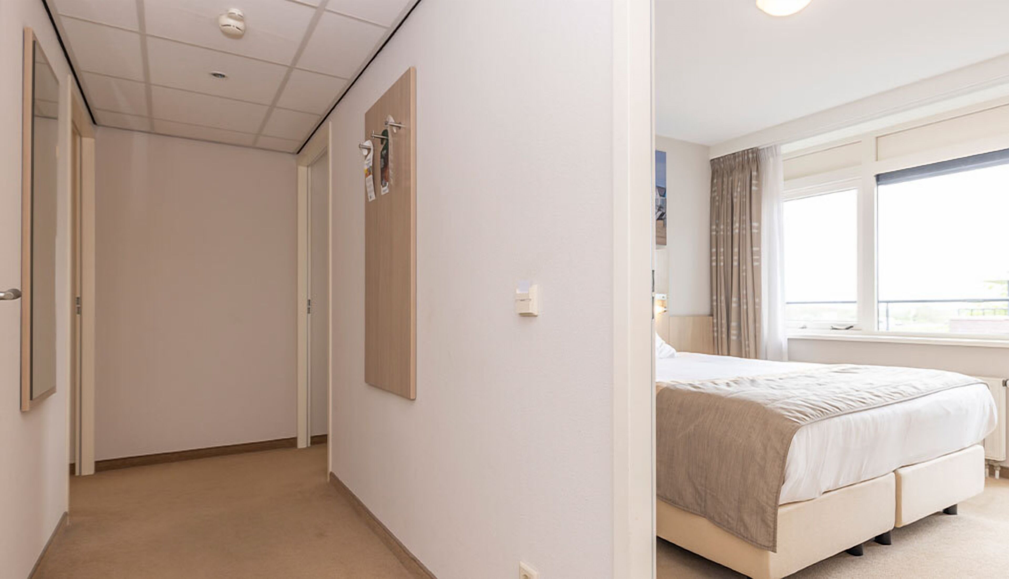 Une chambre d’hôtel avec un lit, une fenêtre avec des rideaux, une porte et un luminaire fixé au mur.