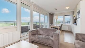 A modern hotel room with large windows, a balcony, a dining area, and a kitchenette.
