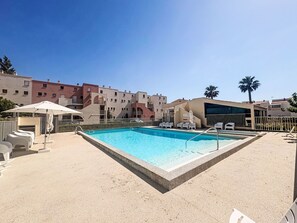 Ein Schwimmbecken mit Liegestühlen, einer überdachten Sitzgelegenheit und einem Gebäude mit Balkonen im Hintergrund.