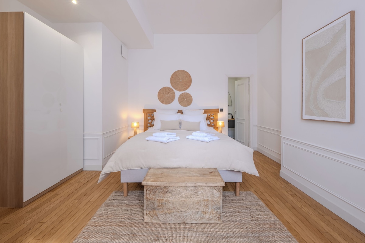 Quarto com cama grande, luminárias de cabeceira, um banco de madeira e uma obra de arte pendurada na parede.