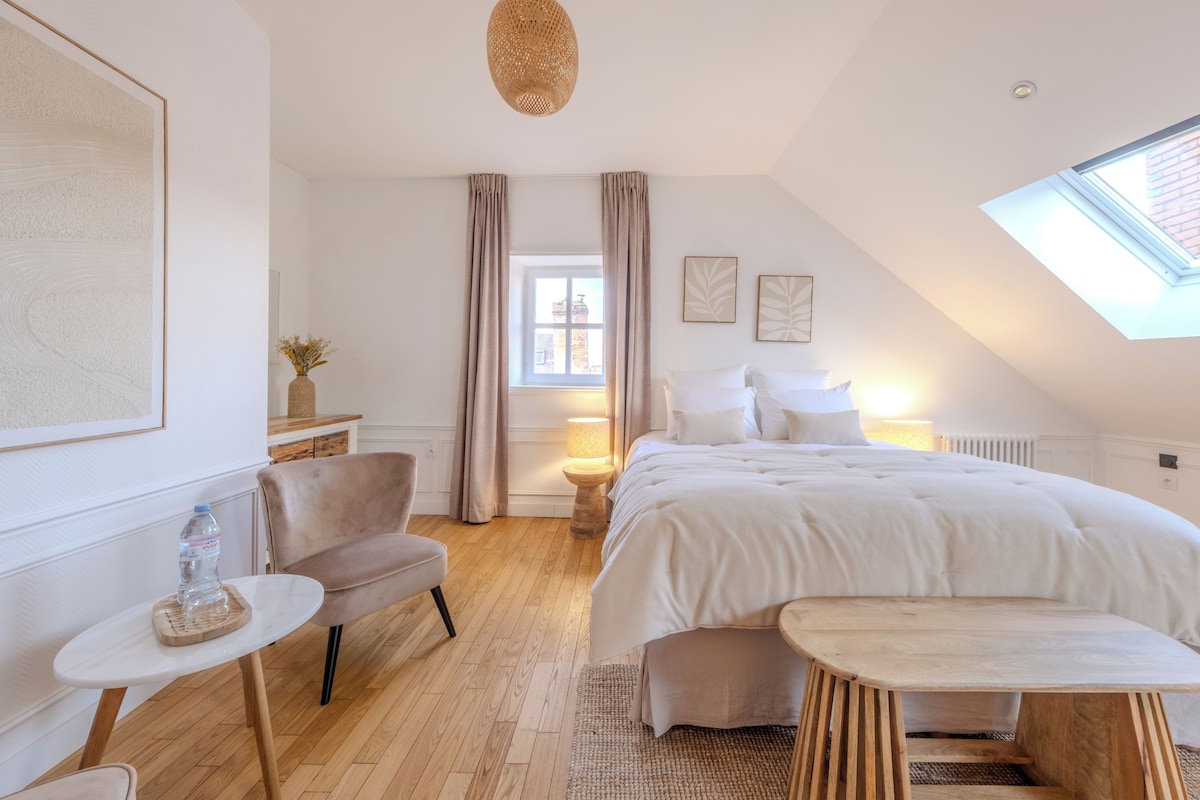 Quarto com cama grande, mesinha, cadeira, janela com cortinas e claraboia.