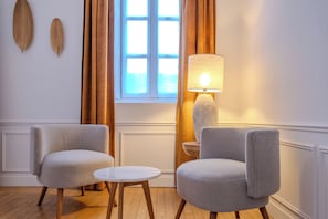 Quarto com duas poltronas, uma mesa redonda pequena, abajur, janela com cortinas e detalhes decorativos na parede.