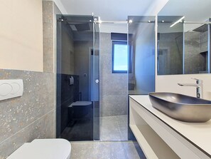 Et moderne badeværelse med brusebad bag glas, et stort spejl og en skålhåndvask.