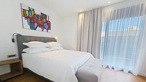 Une chambre moderne avec un grand lit, une table de chevet, une lampe fixée au mur et une fenêtre ornée de rideaux légers.