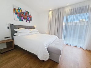 A modern bedroom with a large bed, a bedside table, a wall-mounted lamp, and a window with sheer curtains. - Aloa III by Interhome (Brač/Splitska)