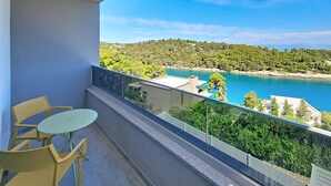 Un balcon avec une table et des chaises, donnant sur un paysage côtier avec des arbres et une étendue d’eau.