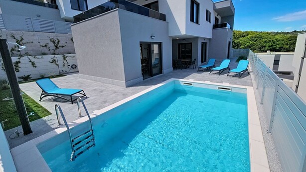 Un espace piscine moderne avec une piscine rectangulaire, des chaises longues et un bâtiment à deux étages doté de balcons.