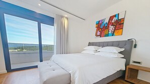 Quarto moderno com uma cama grande, um banquinho, uma mesa de cabeceira, um quadro e vista através de uma janela ampla.