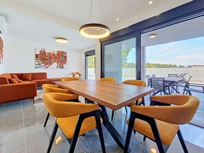 Et moderne spiseområde med et træbord og gule stole, en sofa og et stort vindue med udsigt til naturen.