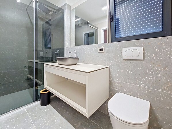 Un baño moderno con cabina de ducha de vidrio, un lavabo blanco con un recipiente, un inodoro y un espejo sobre el lavabo.