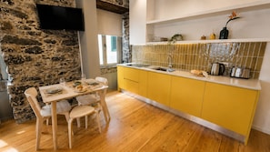 A modern kitchen with a stone wall, wooden dining table, yellow cabinets, and a flat-screen TV mounted above the counter. - Studio apartment | Prime location (Câmara de Lobos)